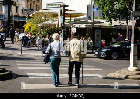 Ein älteres Paar überquert eine Pariser Straße. Die sonnige Szene bietet ein lebhaftes Café im Hintergrund und bietet eine lebhafte Stadtatmosphäre. Id Stockfoto
