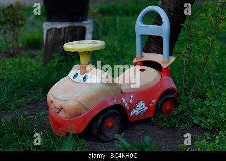 Charmantes altes Spielzeugauto in einem Garten: Ein nostalgischer Anblick eines bunten Kunststoff-Aufsitzfahrzeugs umgeben von grünem Kraut, perfekt für inspirierende Kindheit Stockfoto