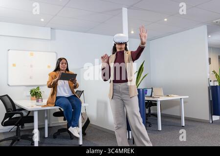 Verschiedene weibliche Kollegen, die VR-Headset und Tablet-PC im Großraumbüro mit Laptops und Pflanzen verwenden Stockfoto