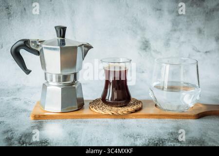 Klassische Mokakanne, frisch gebrühter schwarzer Kaffee in einem schlanken Glas und klares Wasser in einem Tumbler auf einem Bambusbrett vor hellem Steinhintergrund Stockfoto