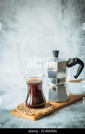 Frischer schwarzer Kaffee in elegantem geschwungenem Glas auf Bambusbrett mit Mokakanne und Wasserglas im Hintergrund, luftig vertikal aufgebautes Layout für Menü oder soc Stockfoto