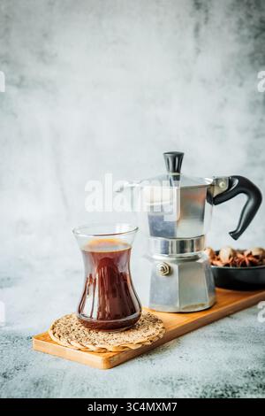Frisch gebrühter Kaffee in schlankem Glas auf Bambusbrett mit Mokakanne und Schüssel mit duftenden Gewürzen sanft verschwimmt dahinter, heller neutraler Hintergrund bereit für Stockfoto