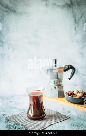 Frisch gebrühter Kaffee in schlankem Glas auf Bambusbrett mit Mokakanne und Schüssel mit duftenden Gewürzen sanft verschwimmt dahinter, heller neutraler Hintergrund bereit für Stockfoto