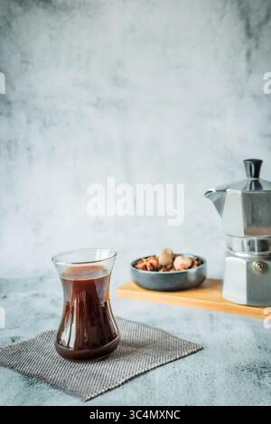 Minimalistisch vertikal mit schwarzem Kaffee in geschwungenem Glas im Vordergrund und verschwommenem Mokakopf mit Gewürzschale dahinter, riesiger Negativraum ideal für Magazin C Stockfoto