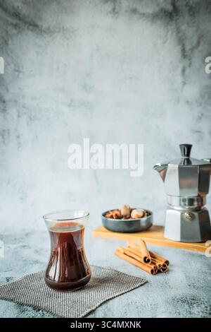 Minimalistisch vertikal mit schwarzem Kaffee in geschwungenem Glas im Vordergrund und verschwommenem Mokakopf mit Gewürzschale dahinter, riesiger Negativraum ideal für Magazin C Stockfoto