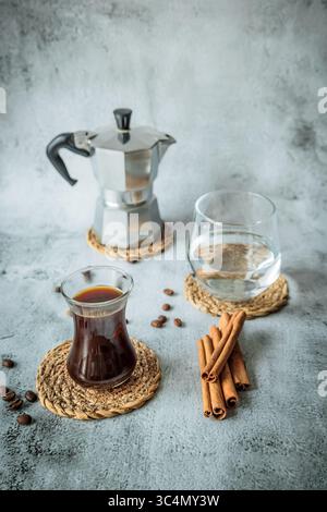 Schwarzer Kaffee in geschwungenem Glas mit Wasserbecher, Mokakanne und Zimtstangen auf gewebten Untersetzern, verstreute Bohnen auf hellem Steinhintergrund, luftig vertikal Stockfoto