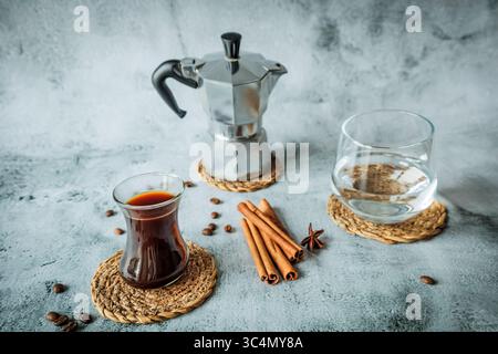 Schwarzer Kaffee in geschwungenem Glas mit Wasserbecher, Mokakanne und Zimtstangen auf gewebten Untersetzern, verstreute Bohnen auf hellem Steinhintergrund, luftig vertikal Stockfoto