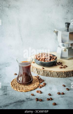 Geschwungenes Glas heißer Kaffee auf der Achterbahn mit verstreuten Bohnen, Schüssel mit gerösteten Bohnen und verschwommenem Mokakopf auf einem Brett hinter dem Baum, geräumige Kulisse für Stockfoto