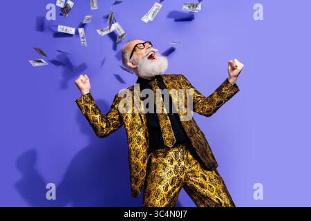 Energiegeladener Seniorenmann in einem stilvollen goldenen Schlangenhautanzug, der vor einem leuchtend violetten Hintergrund aufregend feiert Stockfoto