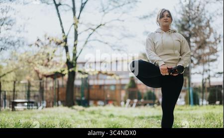 Eine junge Frau, die sich im Park im Freien ausdehnt, in lässiger Trainingskleidung Stockfoto