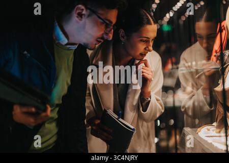 Zwei Personen, die eine Ausstellung in einer schwach beleuchteten Museumsumgebung sorgfältig untersuchen. Stockfoto