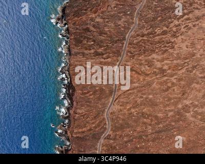 Aus der Vogelperspektive auf die zerklüftete Küste, wo das tiefblaue Meer auf die rötlich-braune Vulkanlandschaft trifft, mit einer Straße, die sich durch das Gelände schlängelt, El Hierro, Kanarische Inseln, Spanien. Stockfoto