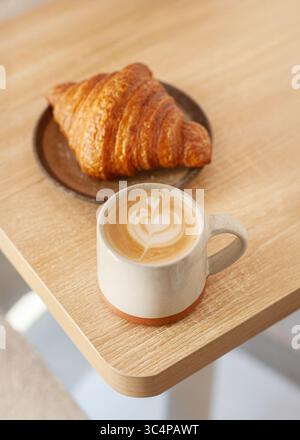 Blick von oben auf ein Croissant auf einer Keramikplatte, serviert mit einer Tasse Cappuccino mit herzförmiger Latte Art. Minimalistisches Frühstück oder Kaffeepausen Stockfoto