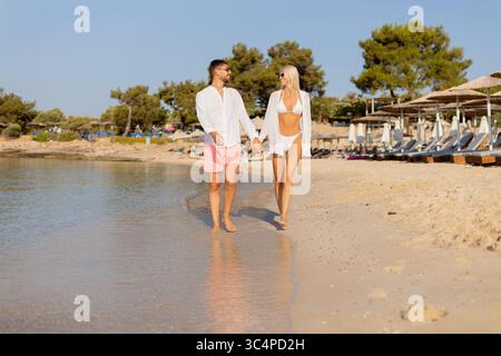 Ein fröhliches Paar geht Hand in Hand am Sandstrand entlang und genießt die sanften Wellen und die warme Sonne. Ihr sorgloses Lachen erfüllt die Luft, als sie mich mitnehmen Stockfoto