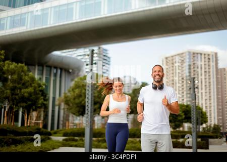 Zwei Freunde machen einen herzerwärmenden Morgenjog durch einen urbanen Park, lächeln und genießen die frische Luft. Die moderne Skyline schafft einen atemberaubenden Hintergrund Stockfoto