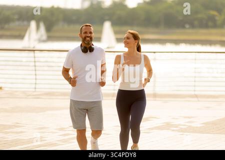 In der warmen Morgensonne joggen ein Paar am Wasser entlang, teilen Lachen und Freude, während sie einen aktiven Lebensstil genießen. Das malerische sce Stockfoto