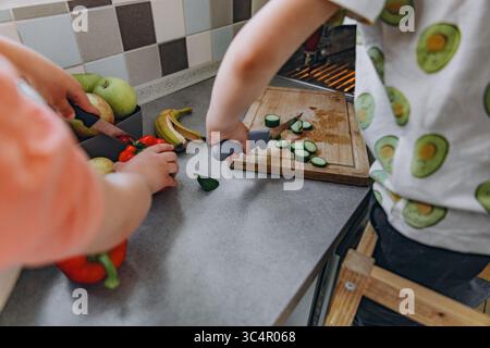 Kinder bereiten Gemüse zusammen in einer hellen Küche zu, schneiden Gurken und genießen die Zeit der Familie. Lernen, Teilen und gesunde Ernährung in A Stockfoto