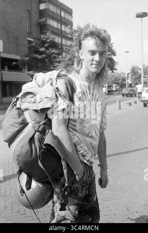 Urlauber NS Station, 07-08-1992 Whizgle News, Dutch Desk, Niederlande, 1950-2000 Ein junger Mann mit einer unverwechselbaren Meeräschen Frisur und einem bunten Tie-Dye Hemd steht selbstbewusst mit einem großen Rucksack über einer Schulter. Er trägt Tarnhosen und einen fröhlichen Ausdruck, der auf Abenteuerlust hindeutet. Die urbane Kulisse bietet eine Mischung aus Gebäuden und Straßenelementen, die auf ein pulsierendes Stadtleben hinweisen. Stockfoto