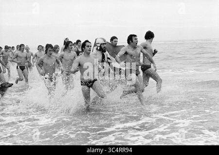 Neujahrstauchgang D.W.R. in Zandvoort, Neujahr, 01-01-1975 Whizgle News, Dutch Desk, Niederlande, 1950-2000 Eine Gruppe von Individuen sprintet in Richtung Ozean und plätschert durch das Wasser, während sie eifrig die Wellen umarmen. Einige Teilnehmer tragen Badeanzüge und zeigen auf dem Weg ins Meer Abenteuerlust und Kameradschaft. Stockfoto