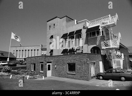 Heliomare Wijk aan Zee, Wijk aan Zee, Relweg, 21.07.1992 Whizgle News, Dutch Desk, Niederlande, 1950-2000 Ein modernes, mehrstöckiges Gebäude verfügt über große Fenster mit Markisen und ein markantes architektonisches Design, das von einer Notentreppe begleitet wird. Im Vordergrund ist eine kleinere Ziegelstruktur zu sehen, in der Nähe befindet sich ein geparktes Auto und eine Flagge, die im Wind winkt. Das Umfeld lässt eine Mischung aus gewerblicher und privater Nutzung in einem städtischen Umfeld vermuten. Stockfoto