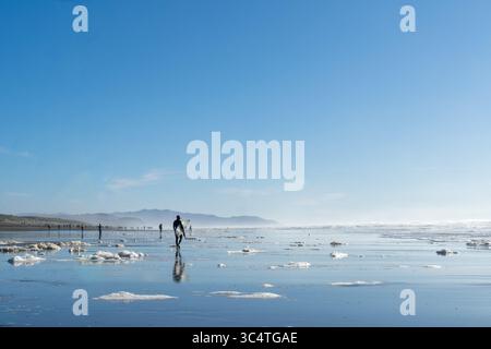 Surfer spazieren am Ocean Beach in San Francisco, Kalifornien, an einem klaren Wintertag mit Meeresschaum und entfernten Silhouetten von Strandgängern. Stockfoto