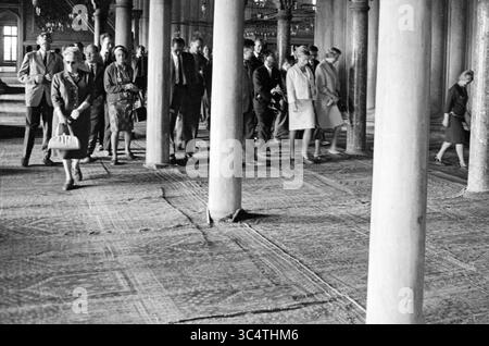 Verkehr und Verschiedenes in Istanbul, Städte, 07-10-1964 Whizgle News, Dutch Desk, Niederlande, 1950-2000 Eine Gruppe von Menschen, die in stilvollen Kleidern aus einer vergangenen Zeit gekleidet sind, schlendern durch ein geräumiges Interieur, das von hohen Säulen gestützt wird. Die Szene vermittelt ein Gefühl der Bewegung und des Zusammenlebens, wobei die einzelnen Personen in Konversation oder Nachdenklichkeit verwickelt sind, während sie durch den gemusterten Boden navigieren. Stockfoto