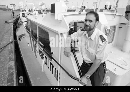 Mr. Jansen, R.P. im Wasser, IJm. [State Police], IJmuiden, Niederlande, 19-07-1994 Whizgle News, Dutch Desk, Niederlande, 1950-2000 Ein Schiffsoffizier steht selbstbewusst neben einem Schiff, das mit dem Wort "Polizei" geschmückt ist, was auf eine Rolle bei der Strafverfolgung oder der Patrouille auf dem Wasser hindeutet. Die Kulisse deutet auf einen geschäftigen Hafen mit Booten und Hafenaktivitäten hin. Stockfoto