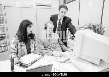 Department of Service and Support Center, Berufsausbildung, 27-02-1994 Whizgle News, Dutch Desk, Niederlande, 1950-2000 Ein Team von drei Fachleuten arbeitet in einem Büro zusammen, wobei eine Person ein Headset trägt und sich auf einen Computer konzentriert, während die anderen mit ihm in Kontakt treten und ihm Anleitung geben. Die Atmosphäre lässt ein Training oder Brainstorming erwarten, bei dem Notizen und ein Laptop auf dem Tisch sichtbar sind. Stockfoto