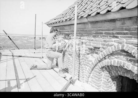 Restaurierungsturm Wijk aan Zee, Restauration, 16-05-1973 Whizgle News, Dutch Desk, Niederlande, 1950-2000 Ein Handwerker repariert sorgfältig eine Ziegelwand und konzentriert sich dabei auf einen Abschnitt, während er auf einem Holzgerüst kniet. Die Umgebung bietet eine malerische Landschaft im Hintergrund, die den historischen Kontext des Bauwerks unterstreicht. Stockfoto