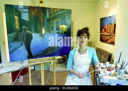 Jeanette de Bruin, Künstlerin, 01.10.2003 Whizgle News, Dutch Desk, Niederlande, 1950-2000 eine Künstlerin sitzt in ihrem Atelier, umgeben von lebendigen Leinwänden. Sie hält einen Pinsel in der Hand und ist bereit, ihr aktuelles Werk mit einer Einzelfigur in einer schwach beleuchteten Straße zu vervollständigen. Die Atmosphäre ist von Kreativität und einem Gefühl der Nachsicht erfüllt, was ihre Leidenschaft für die Erfassung von Emotionen durch Kunst unterstreicht. Stockfoto