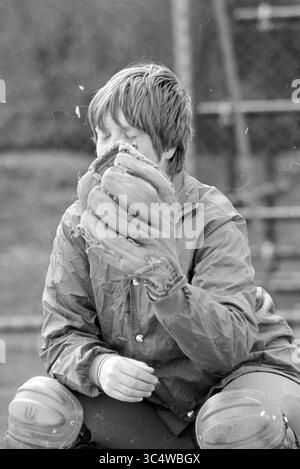Softball-Anschluss. DSC 74 MEN Kennemerland MEN Bloemendaal Women, Softball, 19-04-1980 Whizgle News, Dutch Desk, Niederlande, 1950-2000 Ein junger Mensch sitzt mit einem großen Baseballhandschuh, seine Augen sind teilweise verborgen, während er sich auf ein Spiel vorbereitet. Sie tragen eine leichte Jacke und Fänger-Ausrüstung, die Fokus und Entschlossenheit ausstrahlen, während sie von einer lässigen Sportumgebung umgeben sind. Stockfoto