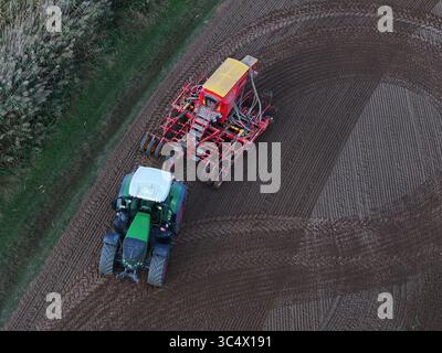 Luftaufnahme eines grünen Traktors, der eine rote Sämaschine über die bearbeitete Erde zieht und gekrümmte Spuren im Boden hinterlässt, Bralitz, Brandenburg, Deutschland. Stockfoto