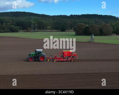 Aus der Vogelperspektive eines grünen Traktors, der eine rote Sämaschine über die frisch bearbeitete Erde zieht, im Kontrast zu den grünen Feldern dahinter, Bralitz, Brandenburg, Deutschland. Stockfoto