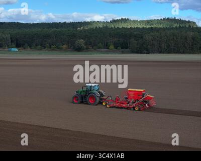 Luftaufnahme eines Traktors mit einer roten Sämaschine, die den Boden bearbeitet, mit einer üppigen Waldkulisse unter einem klaren blauen Himmel, Bad Freienwalde, Brandenburg, Deutschland. Stockfoto