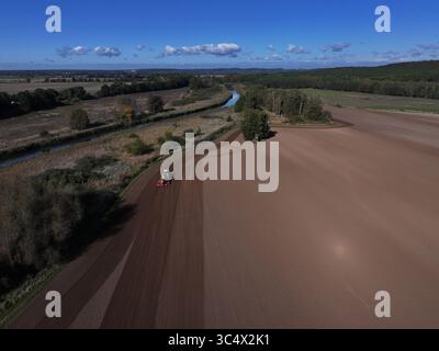 Aus der Vogelperspektive eines Traktors, der die reiche, braune Erde neben einem ruhigen Fluss bebaut, im Kontrast zum grünen Wald unter einem hellblauen Himmel, Bralitz, Brandenburg, Deutschland. Stockfoto