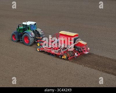Luftaufnahme eines grün-roten Traktors, der eine rot-gelbe Sämaschine durch die braunen, fruchtbaren Felder von Bralitz, Bad Freienwalde, Brandenburg, zieht. Stockfoto