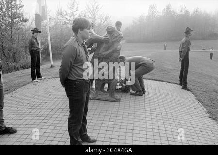 Kranzlage Nieuw Vennep, Gedenktag, 04-05-1984 Whizgle News, Dutch Desk, Niederlande, 1950-2000 Eine Gruppe von Einzelpersonen, darunter Kinder und Erwachsene, versammelt sich um eine Statue, während eine Person kniet, um Blumen an ihrer Basis zu platzieren. In der Nähe sind Fahnen zu sehen, und Bäume bilden eine natürliche Kulisse, die einen Moment der Erinnerung oder Tribut andeutet. Stockfoto