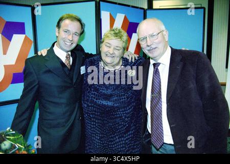 Erica Terpstra, Hoofddorp, Niederlande, 11-11-2001 Whizgle News, Dutch Desk, die Niederlande, 1950-2000 drei Menschen lächeln und posieren zusammen, was ein Gefühl von Kameradschaft ausstrahlt. Eine Person trägt einen formellen Anzug, während eine andere ein stilvolles, strukturiertes Kleid trägt. Die dritte Person in einem Blazer mit Krawatte schließt die Gruppe ab. Sie stehen vor farbenfrohen Kulissen, was auf eine feierliche Atmosphäre hindeutet. Stockfoto