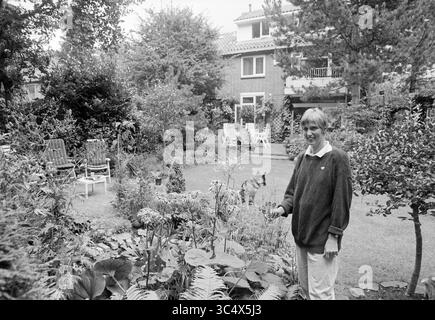 Garden Pride, Els v.d. Struik, A'hout, Aerdenhout, 13-08-1992 Whizgle News, Dutch Desk, Niederlande, 1950-2000 Eine Frau steht inmitten eines üppigen Gartens und lächelt, während sie zu leuchtenden Blumen pflegt. Im Hintergrund befindet sich ein gepflegter Hof mit ordentlich angeordneten Stühlen und einem Blick auf ein gemütliches Zuhause, während ein Hund in der Nähe spielt, was zur ruhigen Atmosphäre beiträgt. Stockfoto