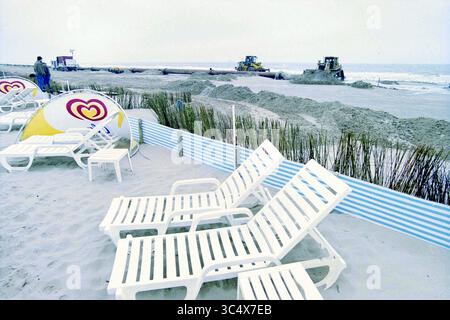Raising the Beach, Z'voort, Zandvoort, 27-05-2001 Whizgle News, Dutch Desk, in den Niederlanden stehen 1950-2000 Liegestühle an einem Sandstrand, in der Nähe arbeiten Maschinen, um die Küste zu Formen. Ein Abschnitt aus blauen Zäunen und Sonnenschirmen verleiht der ruhigen Szene einen Farbtupfer, während die Meereswellen sanft am Ufer herumlaufen. Stockfoto