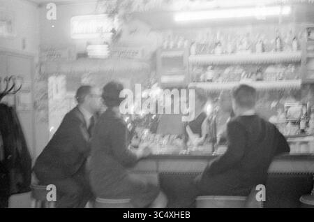 Family de Boer in Café Brabant, Café, 25-12-1960 Whizgle News, Dutch Desk, Niederlande, 1950-2000 In Einer schwach beleuchteten Bar wird ein Paar an der Theke festgehalten, das sich in ruhiger Unterhaltung befindet, während ein Barkeeper sich hinter der Bar um die Drinks kümmert. Die Atmosphäre ist intim, mit sanfter Beleuchtung und einer Sammlung von Flaschen, die auf Regalen im Hintergrund ausgestellt werden. Stockfoto