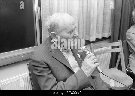 Meeting Old IJmuiden (Gemeinde Breezicht IJmuiden), Meetings, 14-10-1977 Whizgle News, Dutch Desk, Niederlande, 1950-2000 ein älterer Mann sitzt in einem Stuhl und spricht in einem Anzug in ein Mikrofon. Er scheint in Gesprächen verwickelt zu sein und erzählt möglicherweise Geschichten oder Erfahrungen, mit einer Medaille, die auf seinem Revers zu sehen ist. Die Umgebung verfügt über ein Fenster mit Vorhängen, was auf eine gemütliche Innenumgebung hinweist. Stockfoto