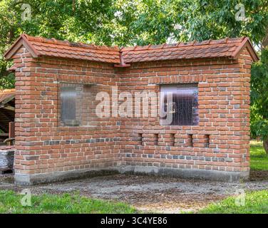 Beltinci, Slowenien - 18. Juli 2025: Ein rustikales Backsteingebäude mit abgeschrägtem Dach, zwei Fenstern und einem einzigartigen Eckdesign. Umgeben von Grün Stockfoto