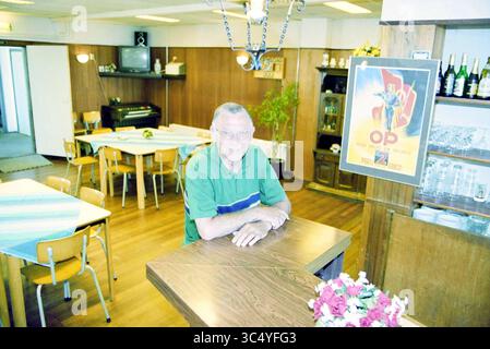 Stadtrat Tjeerd van de Tol, Hillegom, 27-07-1999 Whizgle News, Dutch Desk, Niederlande, 1950-2000 Ein lächelnder Mann mit Brille sitzt an einem Holztisch in einem gemütlichen Essbereich, umgeben von ordentlich angeordneten Stühlen und Tischen. Der Raum verfügt über ein Klavier im Hintergrund, ein dekoratives Poster an der Wand und eine Präsentation von Flaschen, die eine warme und einladende Atmosphäre schaffen. Stockfoto