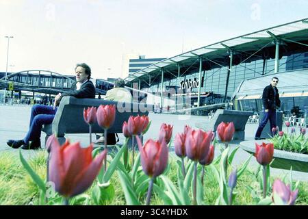Benthem Schiphol, Schiphol, 26-04-2001 Whizgle News, Dutch Desk, Niederlande, 1950-2000 Eine lebhafte Szene auf einem Flughafen mit Tulpen im Vordergrund, während Reisende sich auf Bänken entspannen und eine Person vorbeischlendert und die Mischung aus Natur und Bewegung in einer geschäftigen Umgebung unterstreicht. Stockfoto