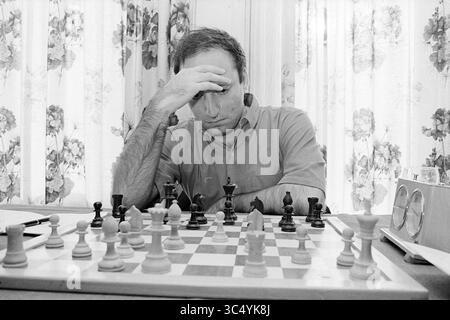 Zuckerman, Blast Furnace Chess Tournament, 15.01.1968 Whizgle News, Dutch Desk, Niederlande, 1950-2000 Ein fokussierter Mensch betrachtet seinen nächsten Zug während eines angespannten Schachspiels, umgeben von einer Reihe von Stücken auf einem karierten Brett. Sonnenlicht zieht durch Blumenvorhänge und verleiht dem strategischen Duell eine ruhige Atmosphäre. Stockfoto
