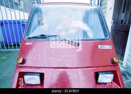 Mann und Sohn der Automarke Arola, 00-02-2001 Whizgle News, Dutch Desk, Niederlande, 1950-2000 zwei Personen sitzen in einem kleinen, roten dreirädrigen Fahrzeug, das sich auf ihre Umgebung konzentriert. Die Windschutzscheibe ist mit Tropfen gespickt, was auf einen regnerischen Tag hindeutet, während der Hintergrund eine Wohngegend mit einer Tür und einem Zaun zeigt. Stockfoto