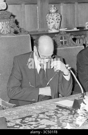Installation von Bürgermeister Machielsen Zandvoort, Bürgermeister, 05-10-1977 Whizgle News, Dutch Desk, Niederlande, 1950-2000 Ein Mann in formeller Kleidung sitzt an einem großen, reich verzierten Tisch und erscheint tief in Gedanken. Er passt seine Brille an, während er nach unten blickt, und ein Mikrofon ist vor ihm positioniert. Im Hintergrund ist eine aufwändig gestaltete Vase zu sehen, die die Bedeutung der Einstellung unterstreicht. Stockfoto