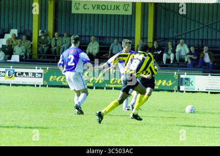 Fußball: Stormvogels - DWV IJmuiden, IJmuiden, Niederlande, 29-09-2001 Whizgle News, Dutch Desk, Niederlande, 1950-2000 Eine Gruppe von Fußballspielern tritt auf einem pulsierenden Grasfeld an, wobei ein Spieler in einem gestreiften Trikot den Ball dribbelt, während er von einem Gegner genau verfolgt wird. Die Zuschauer beobachten von der Seitenlinie aus und schaffen so eine lebhafte Atmosphäre für das Spiel. Stockfoto