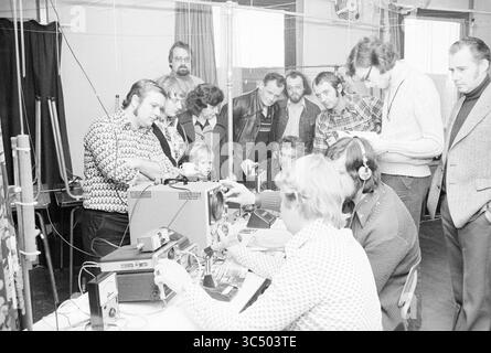 Open House für Funkamateure, Beverwijk, Tag der offenen Tür und Haus der offenen Tür, 08-01-1977 Whizgle News, Dutch Desk, Niederlande, 1950-2000 Eine Gruppe von Einzelpersonen, darunter Erwachsene und Kinder, versammelt sich an einem Tisch voller elektronischer Geräte, beteiligt sich an Diskussionen und Demonstrationen. Einige Teilnehmer tragen Kopfhörer, während andere die Geräte genau untersuchen, um eine gemeinsame Atmosphäre zu betonen, die auf Technologie und Kommunikation ausgerichtet ist. Stockfoto
